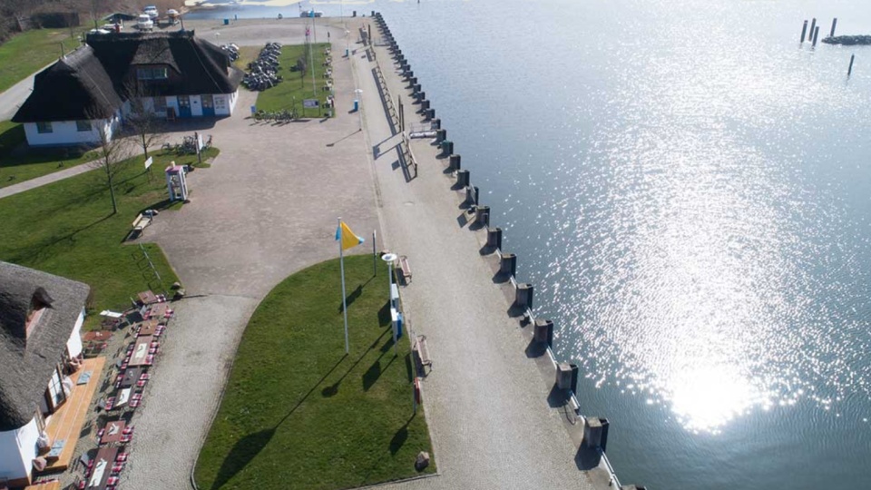 Zu sehen ist eine Hafenmole von oben. Rechts ist das Wasser zu sehen und links auf der Mole erkennt man das Restaurant Hafenamt in Kloster auf der Insel Hiddensee.