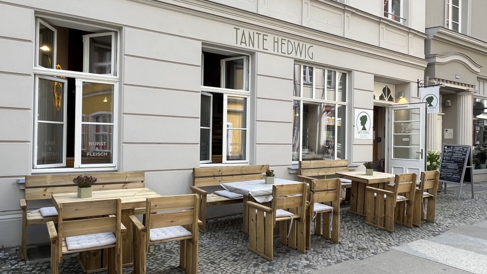 Zu sehen ist die Außenfassade des Hoflandes Tante Hedwig in Stralsund. Vor dem Laden stehen Stühle, Bänke uns Tische aus massivem Holz die zum Verweilen einladen. Der richtige Ort für ein Frühstück, eine Vesper oder nur einen Kaffee.