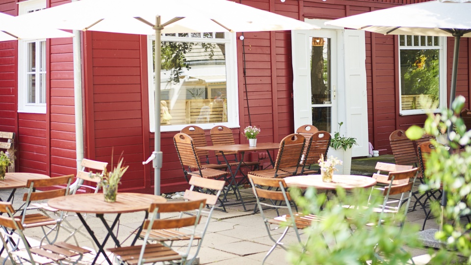 Auf diesem Bild zu sehen ist das Restaurant Das Rote Haus in Vitte auf der Insel Hiddensee von außen. Die Holzfassade ist rot gestrichen, vor dem Haus stehen braune Tische und Stühle unter weißen Sonnenschirmen, die zum Sitzen und Verweilen einladen.