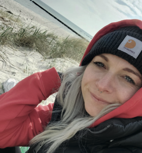 Sabine Dirschke mit Mütze und Jacke am Strand von Hiddensee. Im Hintergrund sieht  man grünes Strandgras und die Ostsee.