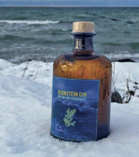 Eine Flasche Bernstein-Gin – Dry Gin von der Insel Hiddensee steht auf dem blauen Etikett –  im Schnee vor bewegter Ostsee auf der Insel Hiddensee